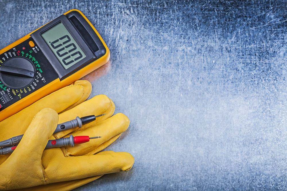 Digital multimeter and leather work gloves with test leads on a scratched metal bench